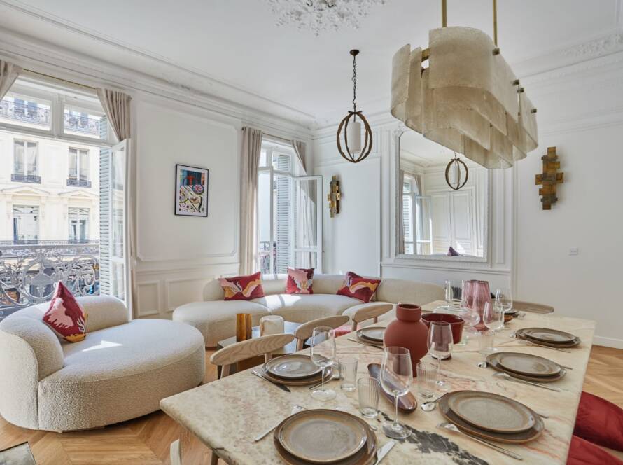 Salle à manger lumineuse avec grande table, assises douces et coussins rouges; fenêtres anciennes et décor blanc élégant évoquant une rénovation complète maison île-de-france.
